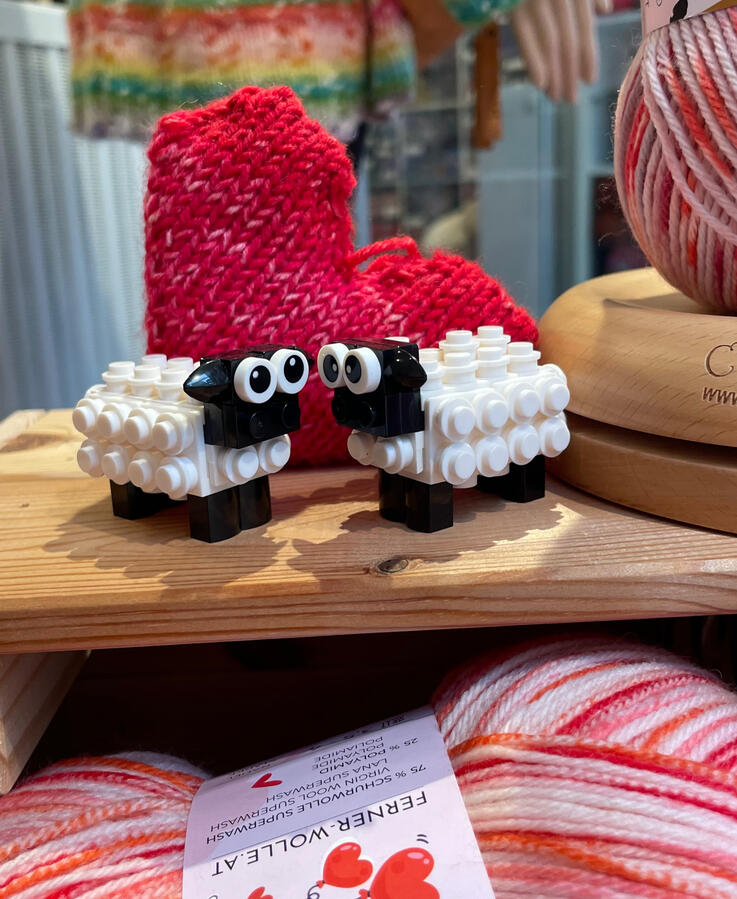 Valentinsschäfchen Zu sehen ist ein Schäfchenpaar aus Klemmbausteinen, der Hintergrund ist dekoriert mit einem gestrickten Herz.